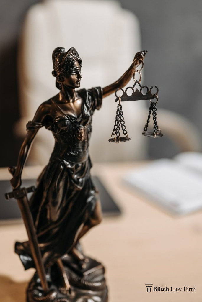 Bronze statue of Lady Justice holding hanging scales in one hand and a sword in the other, with a law firm watermark in the corner.